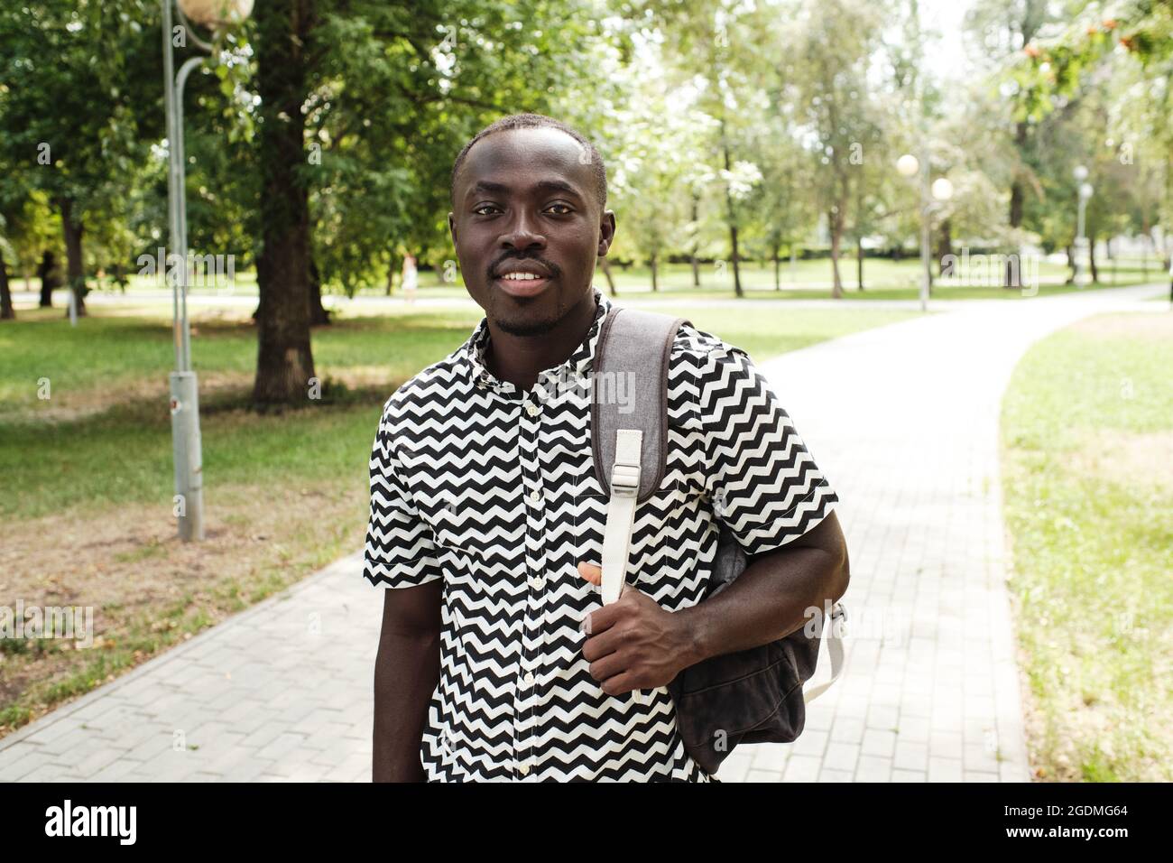 Porträt eines afrikanischen Studenten mit Rucksack hinter seinem Rücken, der die Kamera anlächelt, während er durch den Park geht Stockfoto