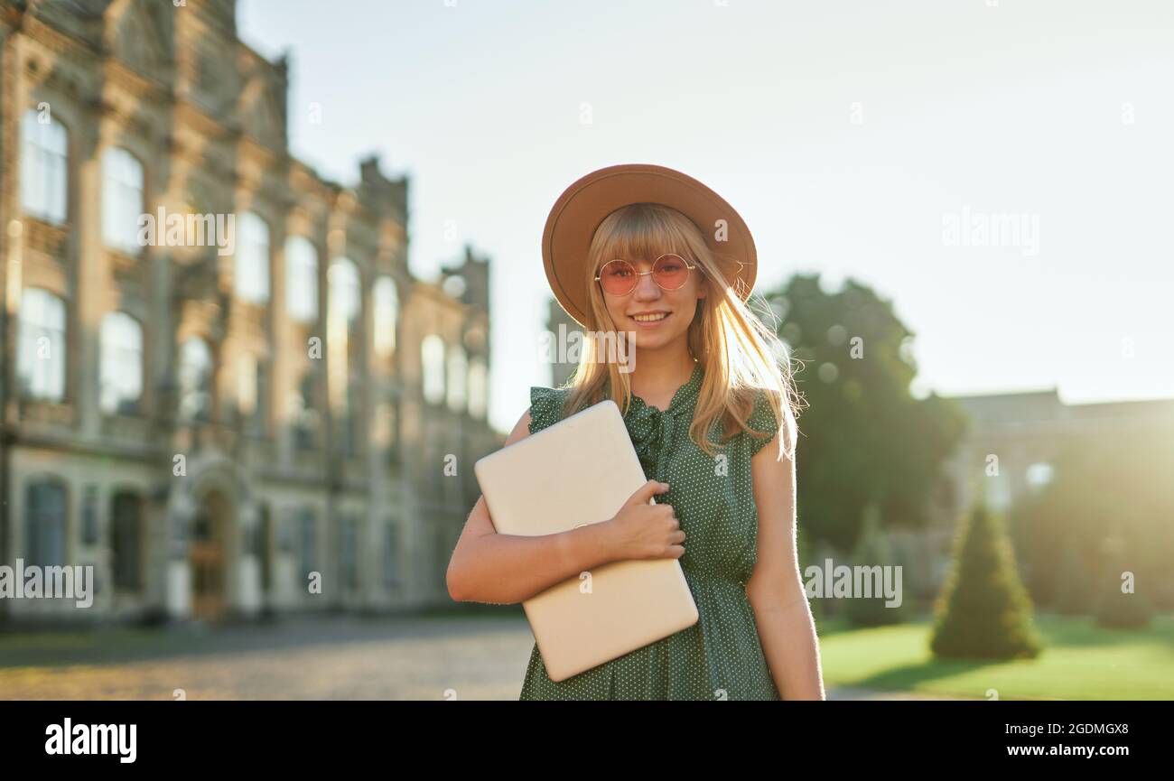 Nettes fröhliches Schulmädchen oder pädagogisches Konzept. Junge blonde Studentin mit Laptop, rosa Sonnenbrille, grünem Kleid und Hut auf dem Universitätscampus. Hochwertige Bilder Stockfoto