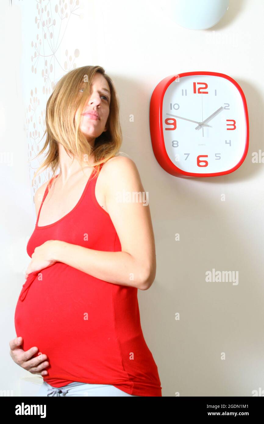 Eine Schwangerin (neun Monate) sieht erwartungsvoll auf eine Uhr. Model Release Verfügbar Stockfoto