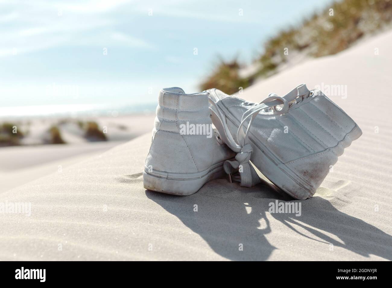 Ein paar weiße Sneakers am Strand auf Sand gelassen. Dünen mit grünem Gras Vegetation. Sommerurlaub, Urlaub am Meer Konzept. Stockfoto