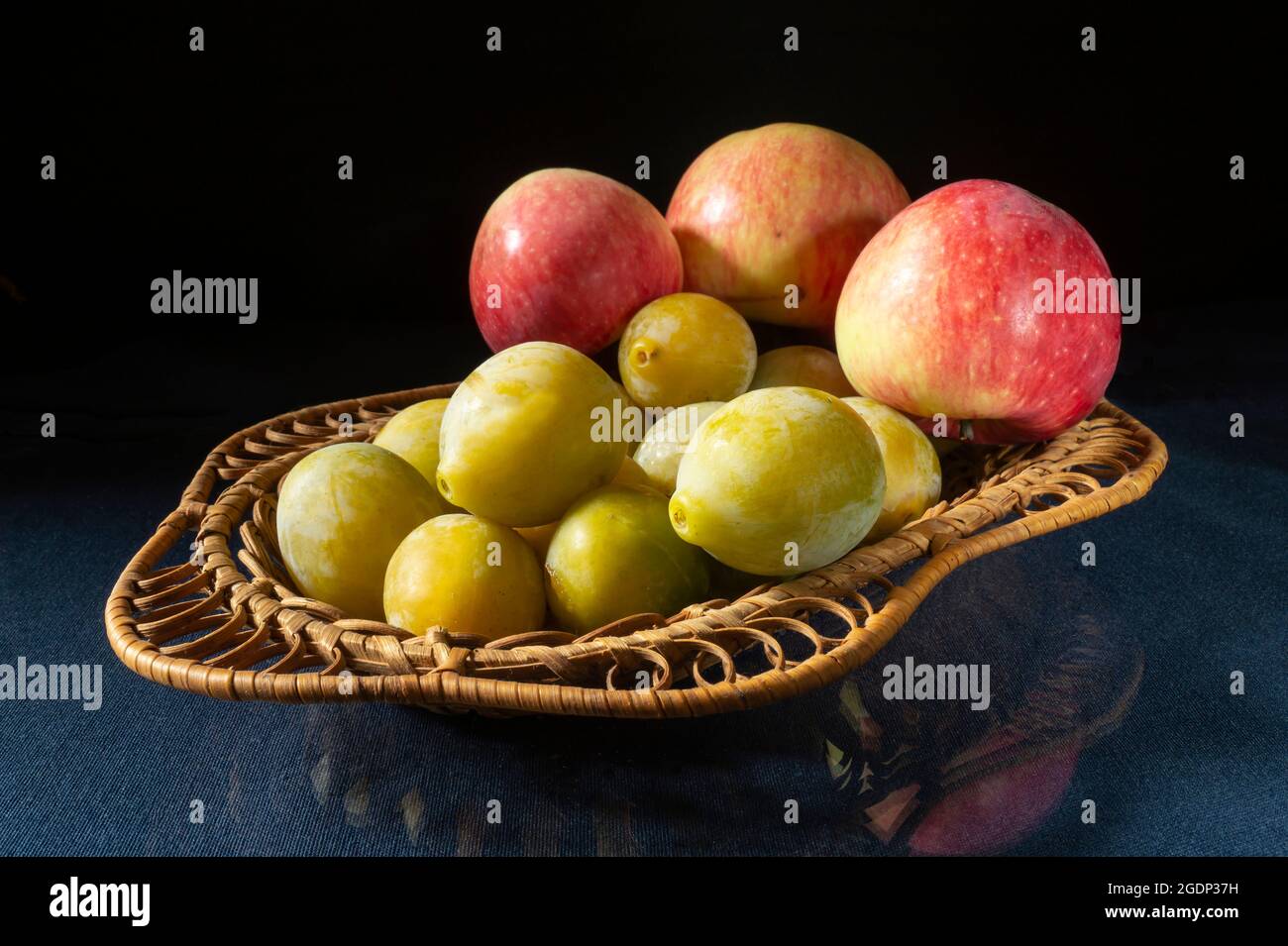 Honigpflaumen mit Äpfeln in einem Korb auf dem Tisch mit einer Reflexion. Lebensmittel auf schwarzem Hintergrund Stockfoto