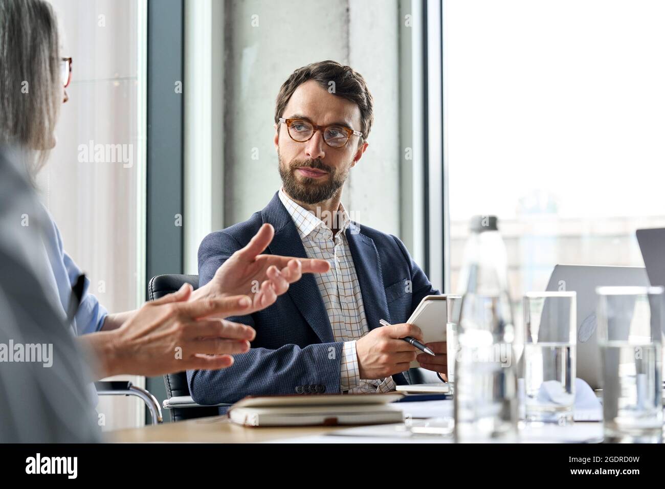 Ernsthafter, männlicher ceo hört einer Kollegin bei der Tischbesprechung zu, wie sie das Notizbuch hält. Stockfoto