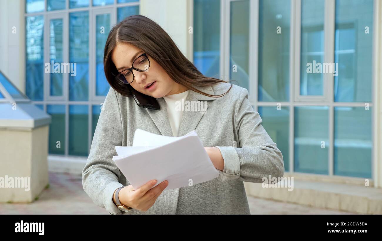 Eine junge Frau in einem formellen Anzug und einer Brille telefoniert und prüft unterwegs Dokumente Stockfoto