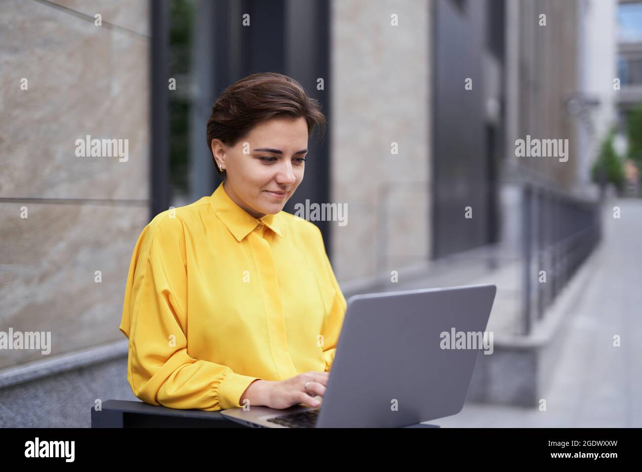 Nette selbstbewusste Brünette Geschäftsfrau in gelber Bluse steht draußen auf der Straße mit einem Laptop arbeiten kaum, tippen. Business, Finanzindustrie oder Immobilienkonzept. Hochwertige Bilder Stockfoto