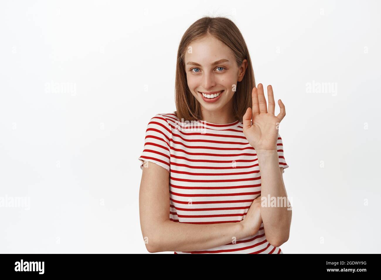 Schöne junge Frau kichert schüchtern, winkt mit der Hand zu Ihnen, sagen Sie Hallo kokett, lächelnd und winken hallo Geste, Gruß jemand, stehend in gestreiftem T-Shirt Stockfoto