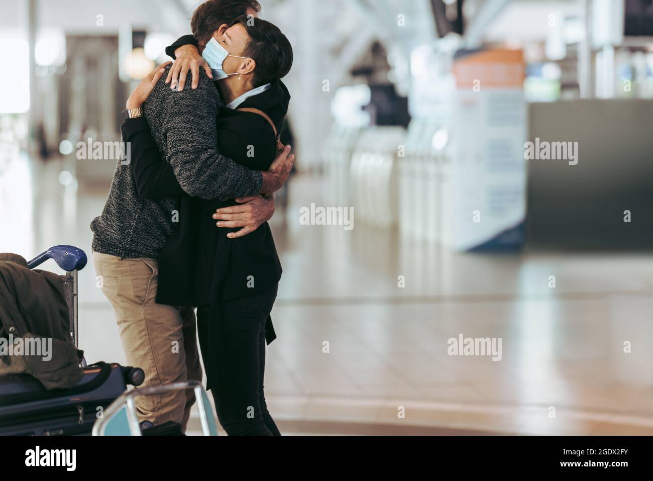 Frau in Gesichtsmaske umarmte Mann am Ankunftseingang des Flughafens. Frau umarmte und begrüßte den Mann nach einer Pandemie am Flughafen. Stockfoto