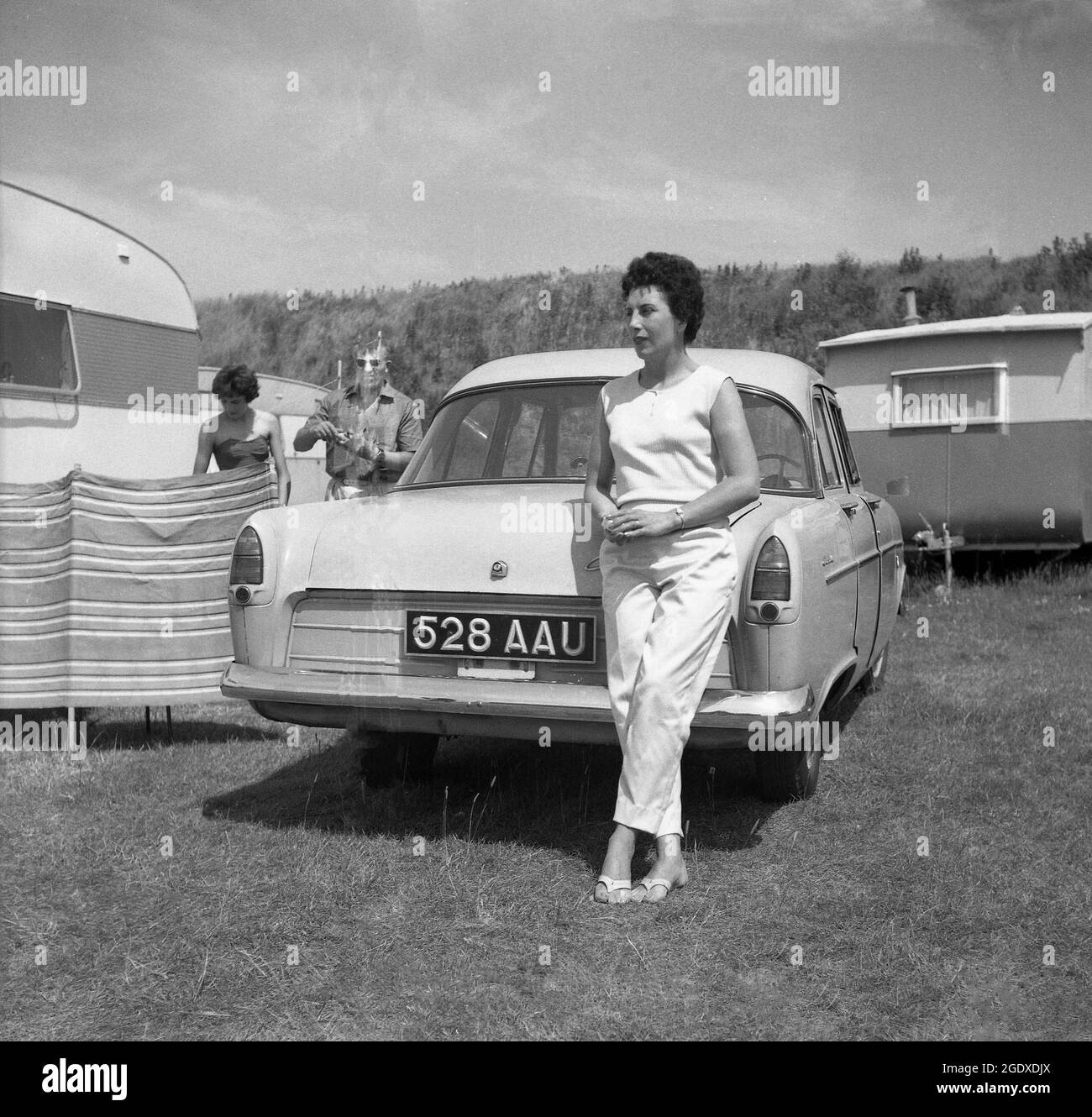 1960s, historisch, draußen auf einem Feld auf einem Caravanplatz, eine stilvolle Dame, die sich auf den Kofferraum eines Autos der Zeit lehnte, neben einem Windbrecher geparkt, neben einem Wohnwagen, England, Großbritannien, aufgestellt. Stockfoto