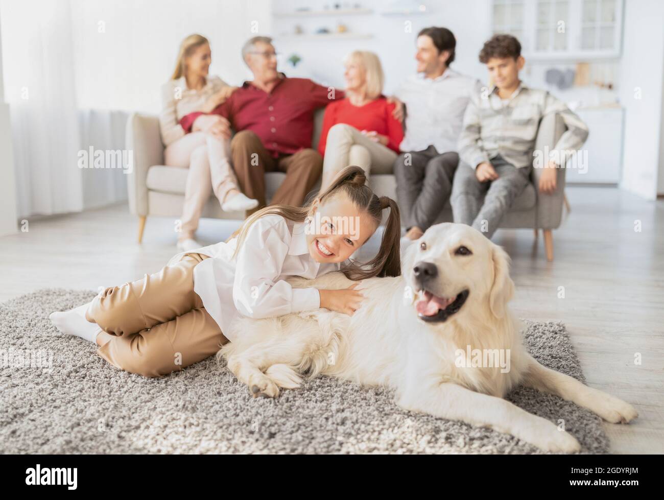 Glückliches Mädchen mit niedlichem Hund, der auf dem Boden liegt, und ihrer Familie mit mehreren Generationen, die auf der Couch im Wohnzimmer miteinander kommunizierte Stockfoto