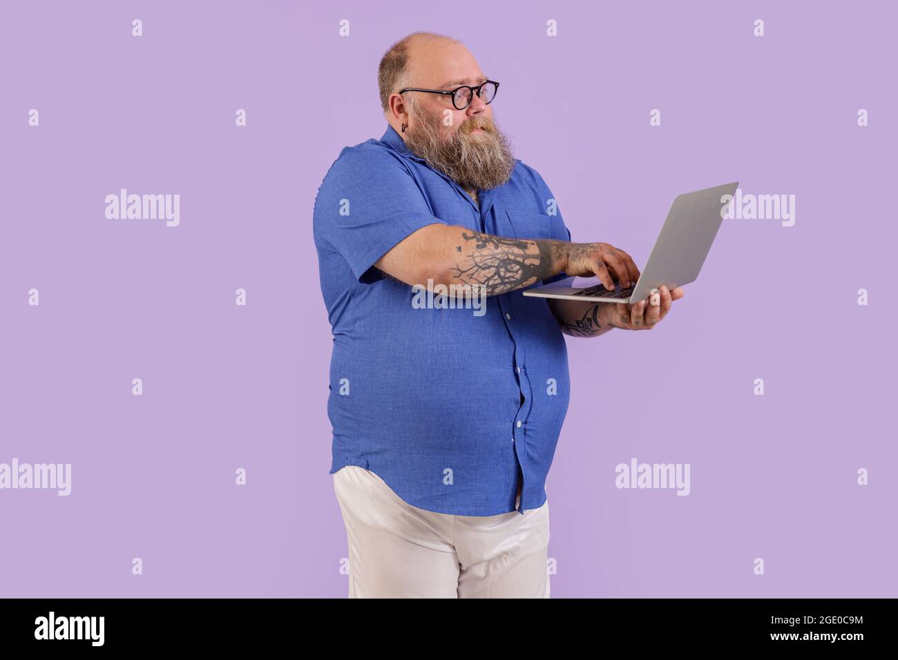 Ein emotionaler Mann mit Übergewicht arbeitet auf einem Laptop, der im Studio auf violettem Hintergrund steht Stockfoto