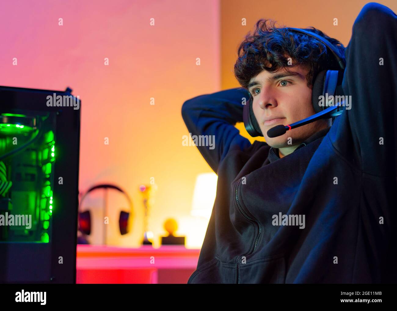 Der junge kaukasische Teenager Gamer mit Kopfhörern lehnt sich im Stuhl zurück, die Arme hinter dem Kopf und das Gesicht sind zufrieden. Stockfoto