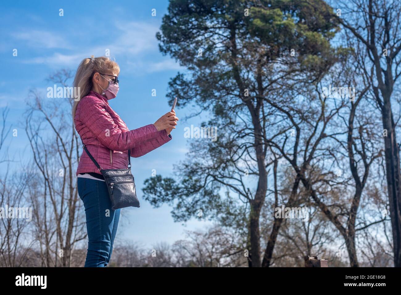 Beschnittene Ansicht einer erwachsenen Frau mit Gesichtsmaske und Sonnenbrille, die ein Foto in einem Park mit einigen Bäumen hinter sich nimmt. Stockfoto