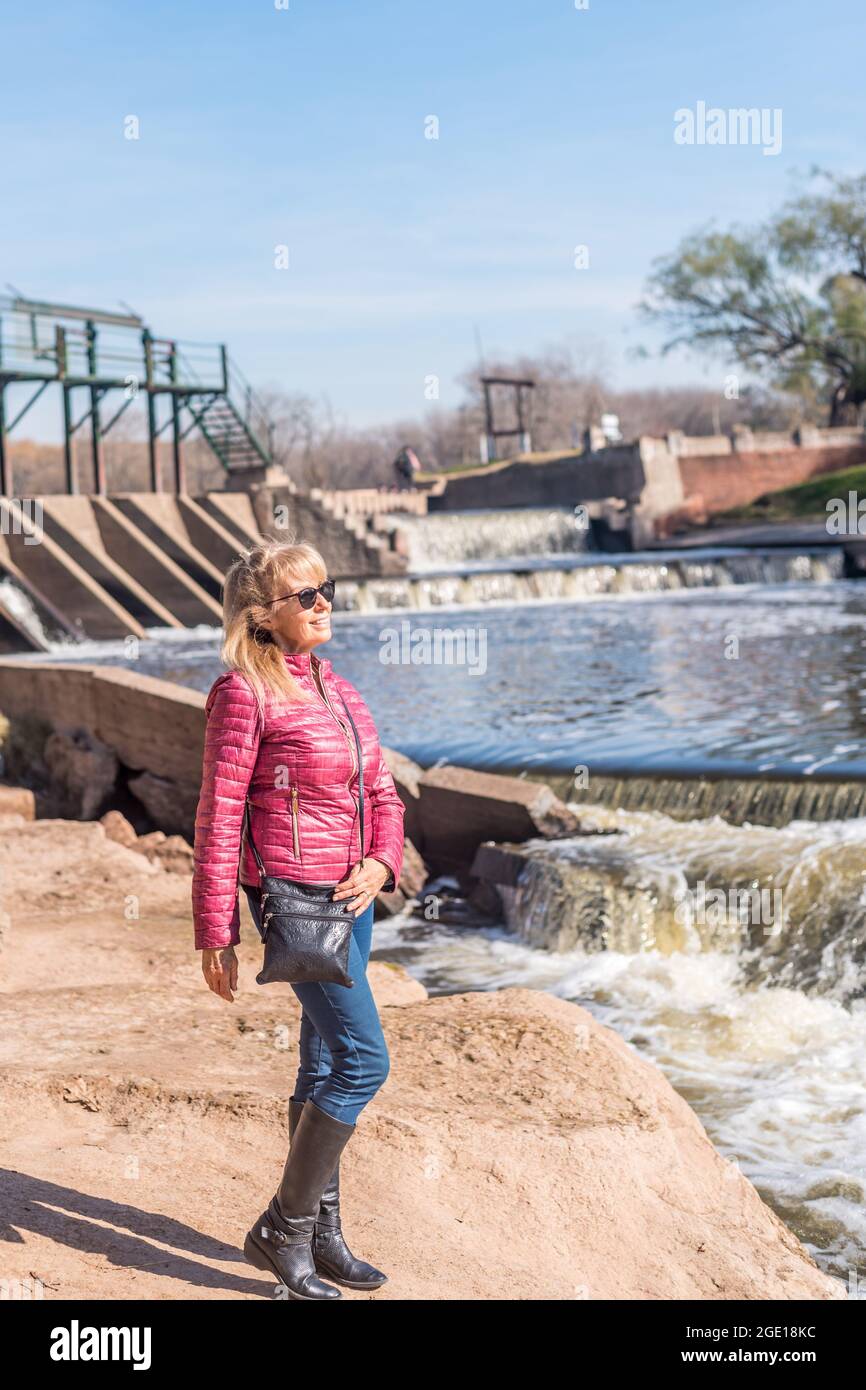 Porträt einer fröhlichen erwachsenen Frau, die an einem sonnigen Tag mit einer Sonnenbrille im Park mit einem Teich hinter sich steht. Stockfoto