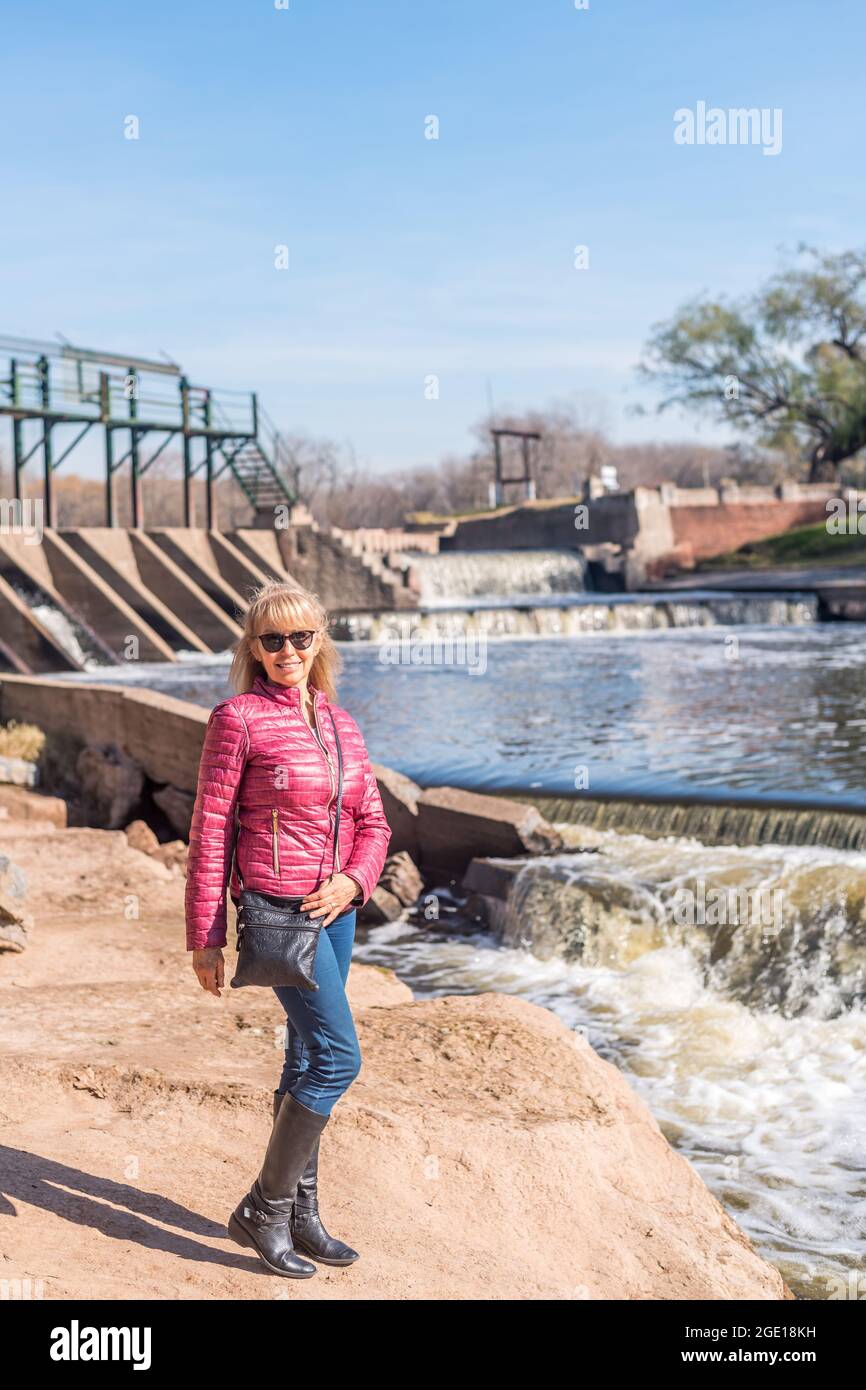 Porträt einer fröhlichen erwachsenen Frau mit Sonnenbrille, die an einem sonnigen Tag im Park mit einem Teich hinter sich steht. Stockfoto