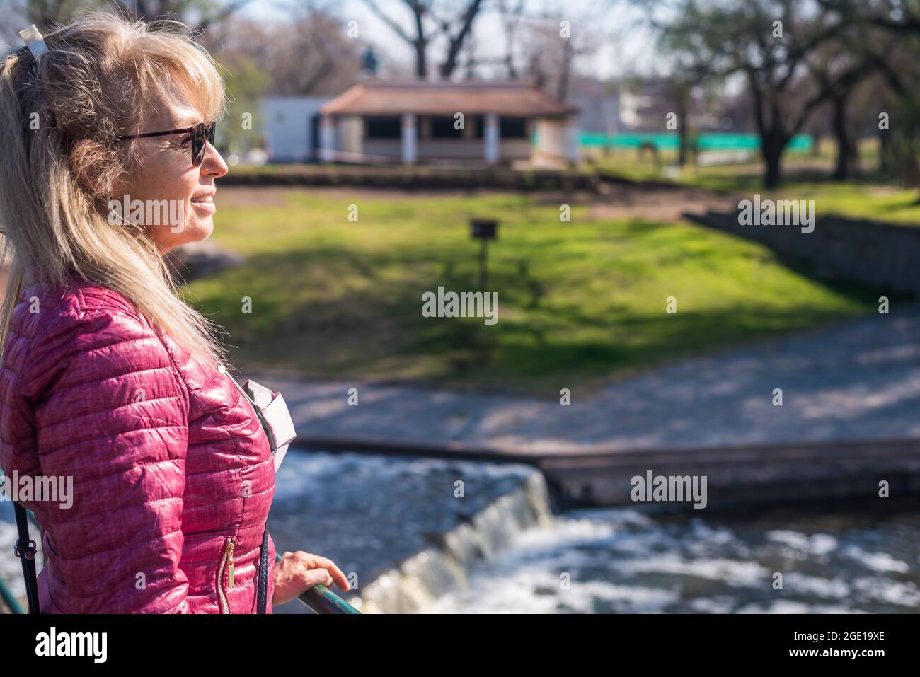 Nahansicht einer fröhlichen erwachsenen Frau mit einer Sonnenbrille, die im Park auf dem Geländer mit einem Teich und grünem Gras hinter sich steht und nach innen schaut Stockfoto