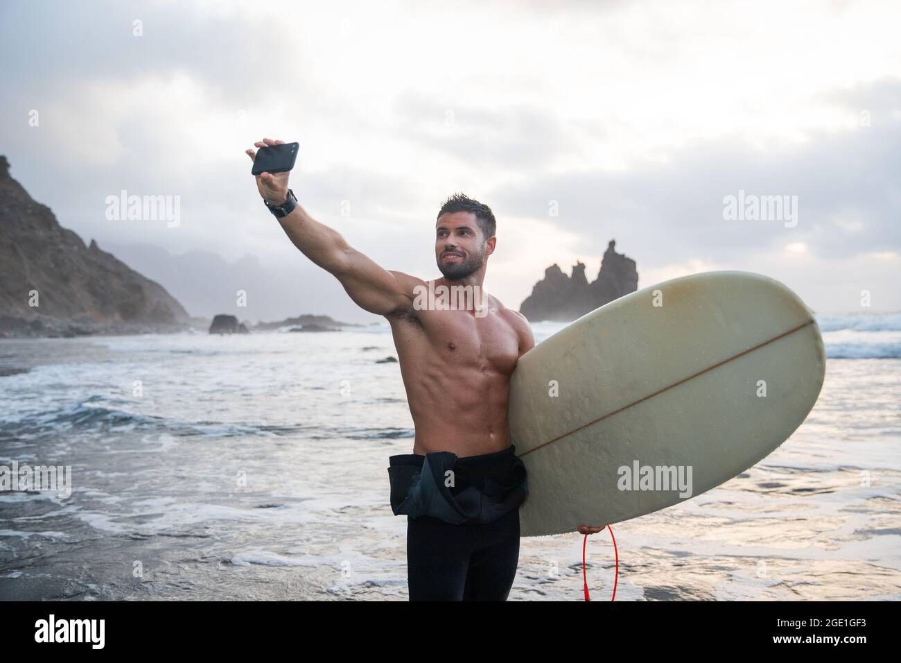 Sportlicher junger Surfer macht ein Selfie am Strand, sportlicher Typ hält ein Surfbrett und steht während des Sonnenuntergangs an einem tropischen Strand - sportlich tausendjährig Stockfoto