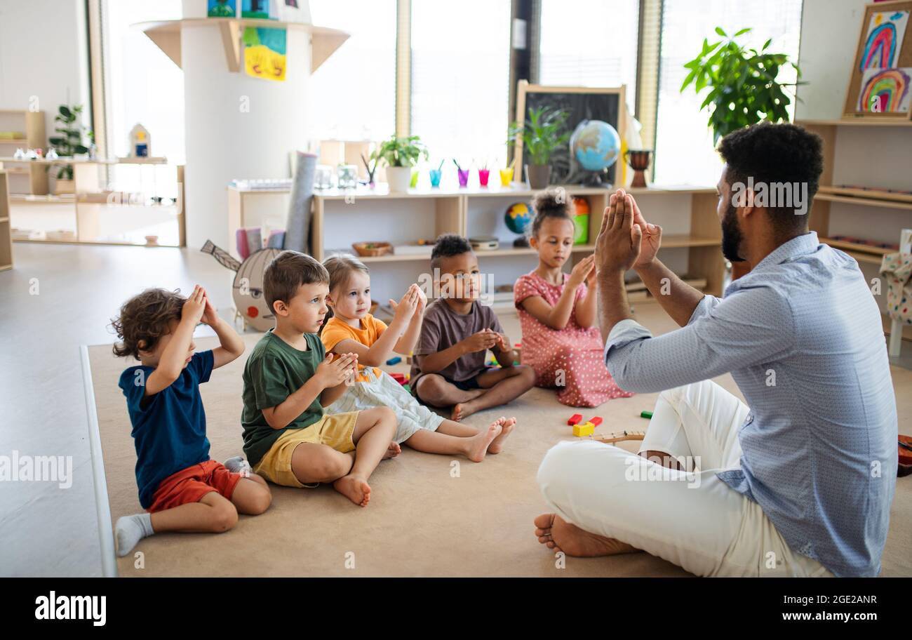 Gruppe von kleinen Kindern im Kindergarten mit einem Lehrer, der im Klassenzimmer auf dem Boden sitzt und spielt. Stockfoto