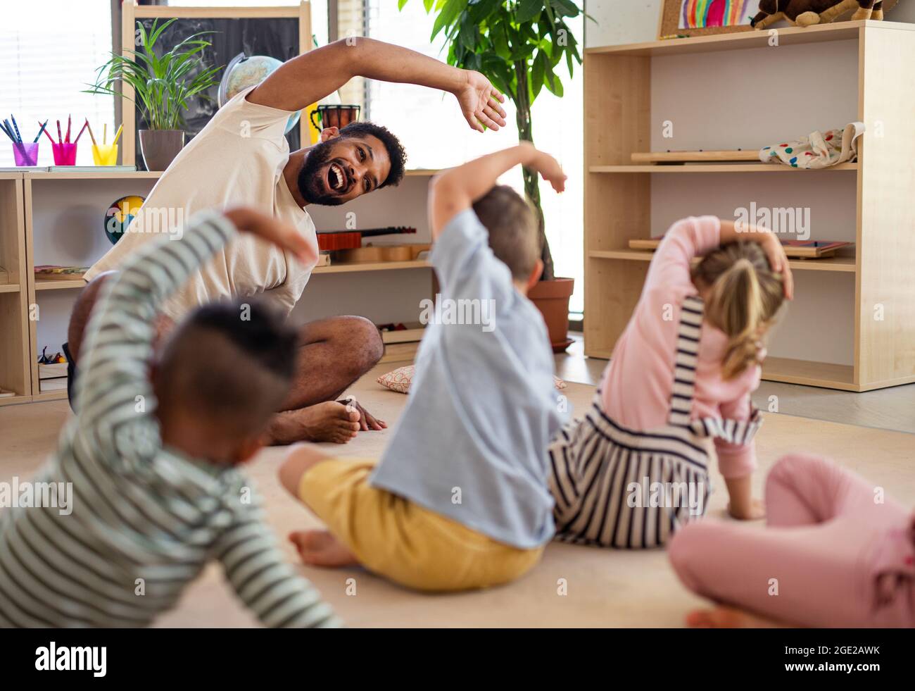 Gruppe von kleinen Kindern im Kindergarten mit einem Lehrer, der im Klassenzimmer auf dem Boden sitzt und Übungen macht. Stockfoto