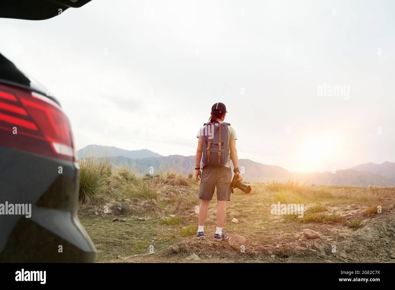 Rückansicht eines asiatischen Fotografen, der mit dem Auto unterwegs ist, mit der Kamera in der Hand und Blick in der Morgensonne Stockfoto