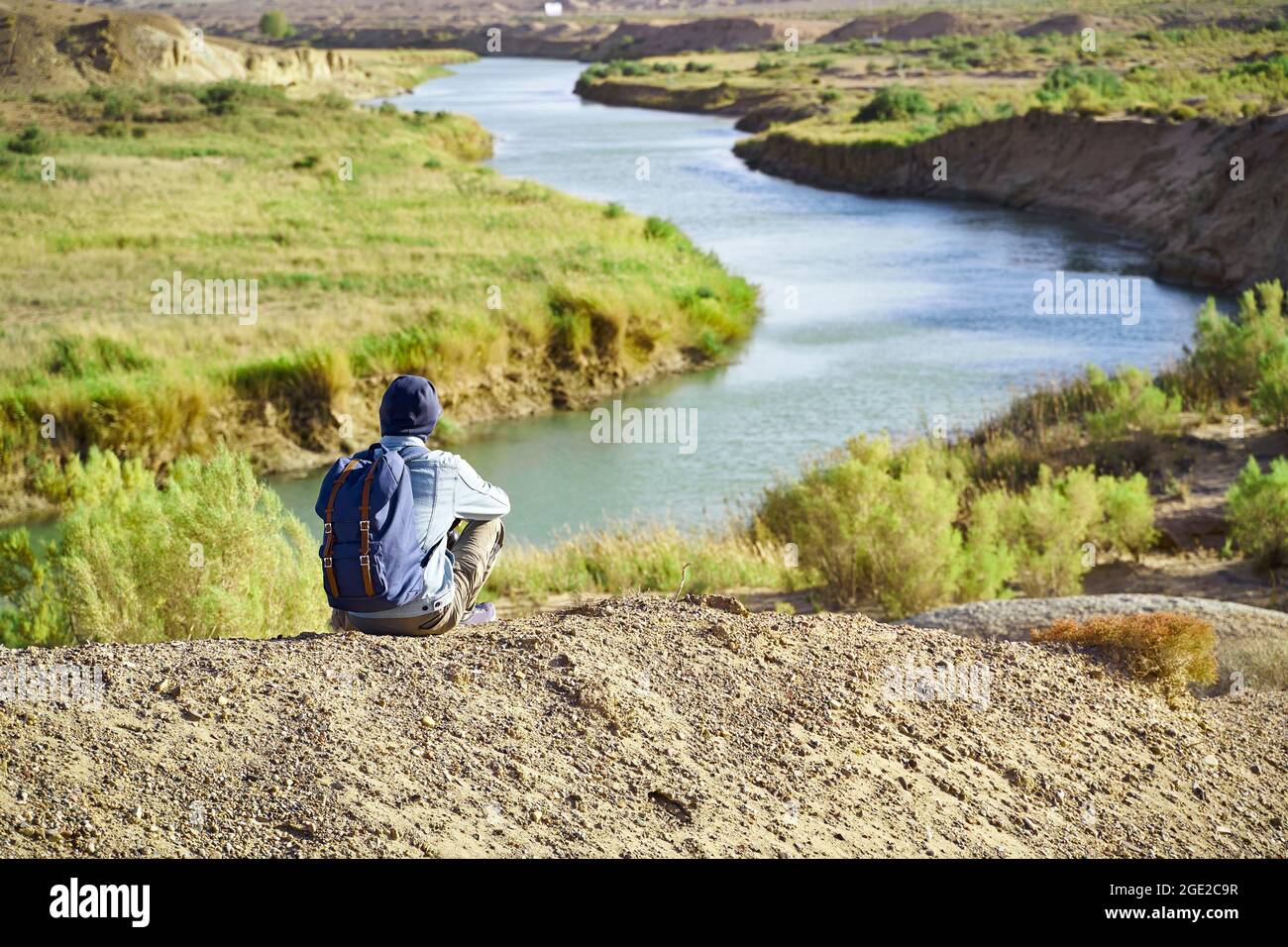 Rückansicht eines männlichen asiatischen Fotografen, der auf einem Hügel sitzt und auf einen Fluss blickt Stockfoto