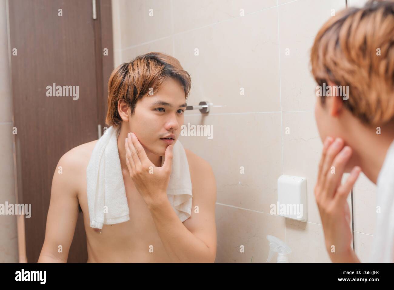 Der junge Mann flattert sein Gesicht und steht mit einem Handtuch auf seinen Schultern vor dem Badezimmerspiegel Stockfoto