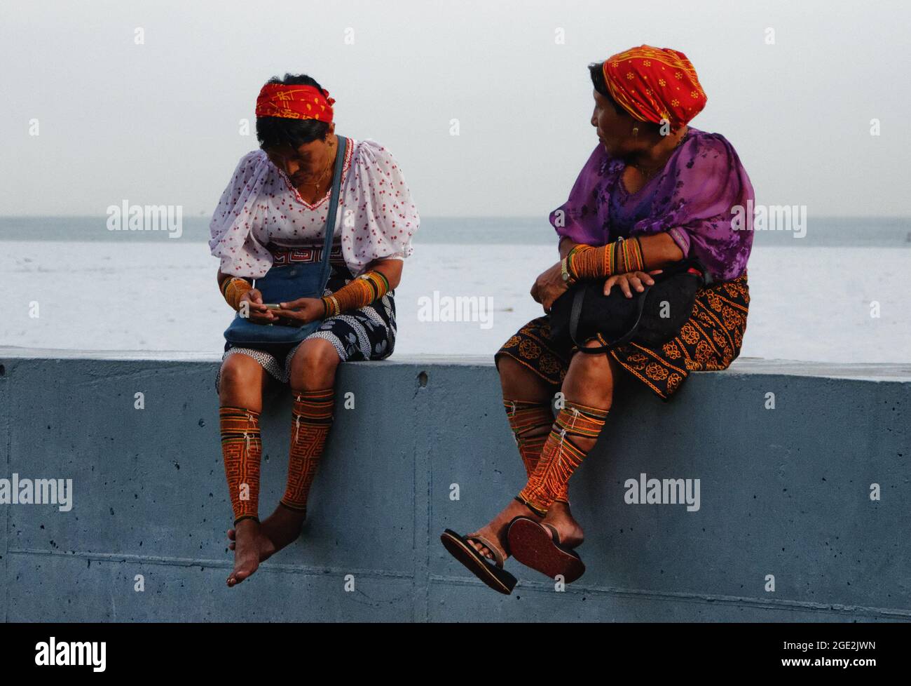 Zwei reife Frauen in traditioneller Kleidung, die sich an der Meeresmauer im Mirador, Coastal Belt, Panama City, ausruhen, eine überprüft ihr Mobiltelefon. Stockfoto