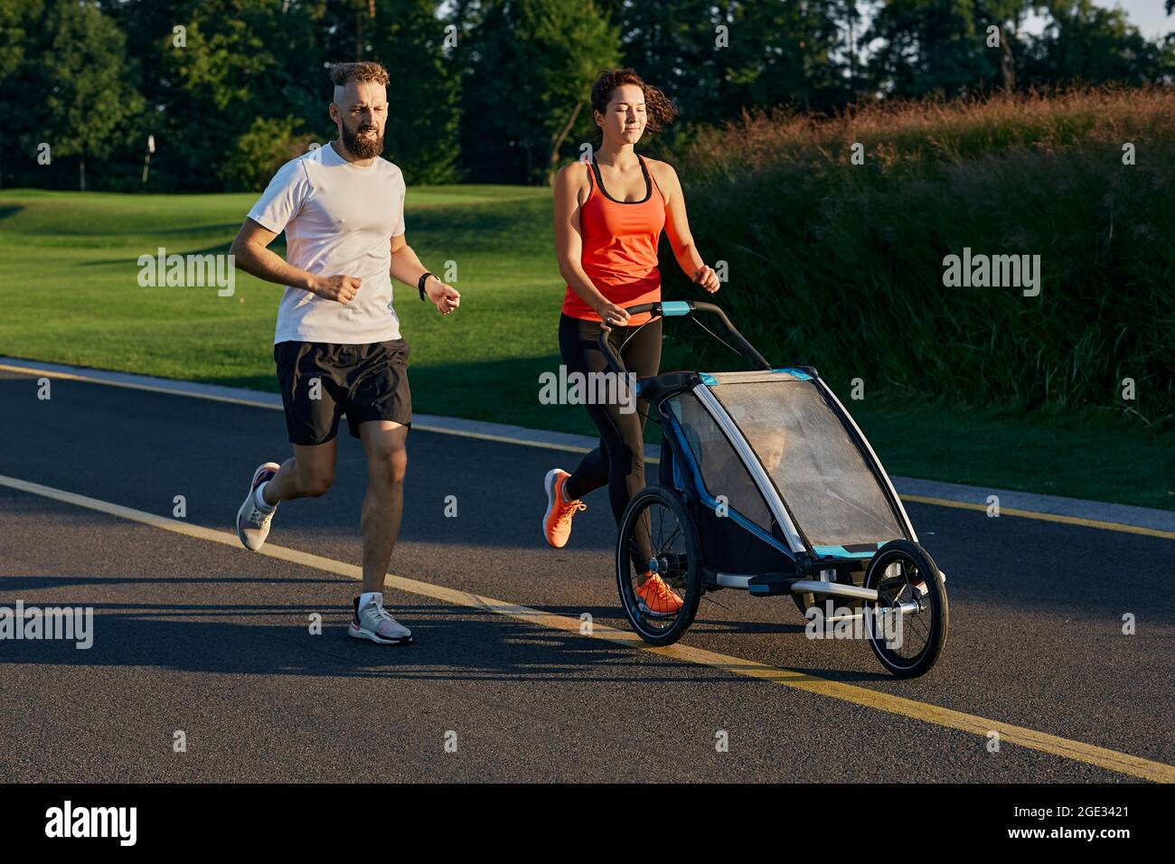 Aktives Familienleben. Junge Familie mit ihrem Kind in einem Jogging-Kinderwagen beim Joggen in einem öffentlichen Park Stockfoto