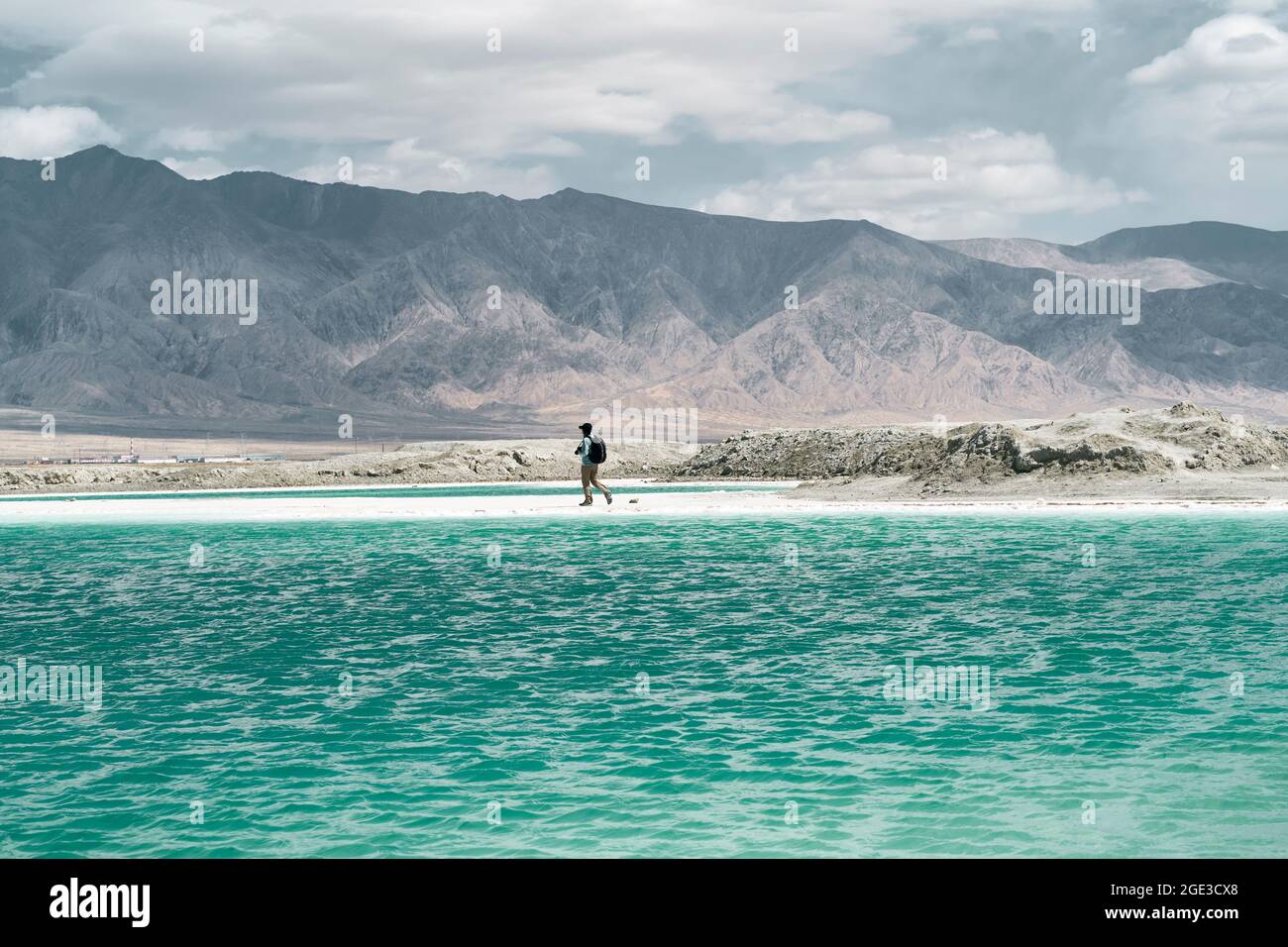 asiatischer Mann, männlicher Fotograf, der an einem Salzsee mit hügeligen Bergen im Hintergrund entlang geht Stockfoto