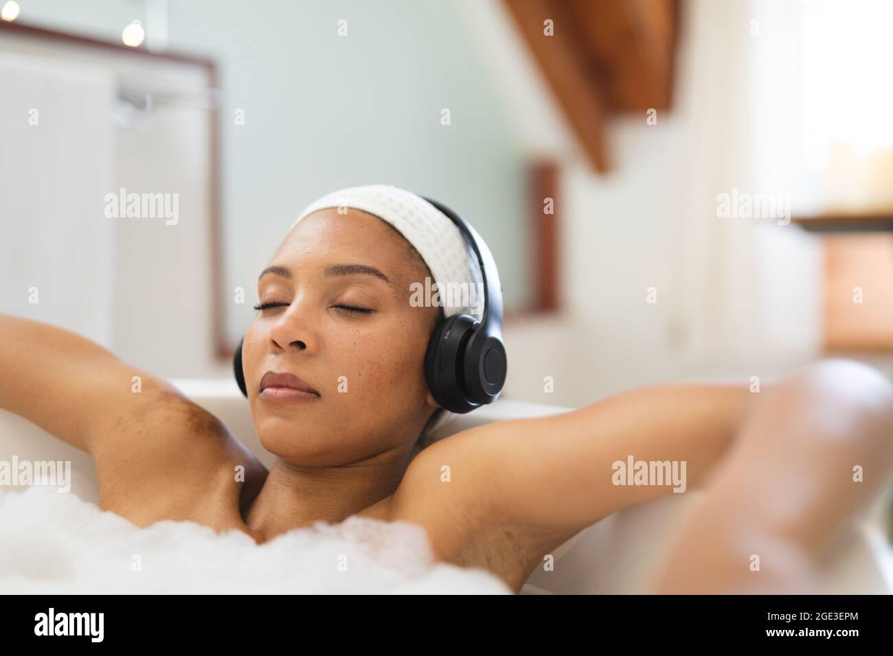 Frau mit gemischter Rasse im Badezimmer, die sich in der Badewanne mit Kopfhörern und geschlossenen Augen entspannt Stockfoto