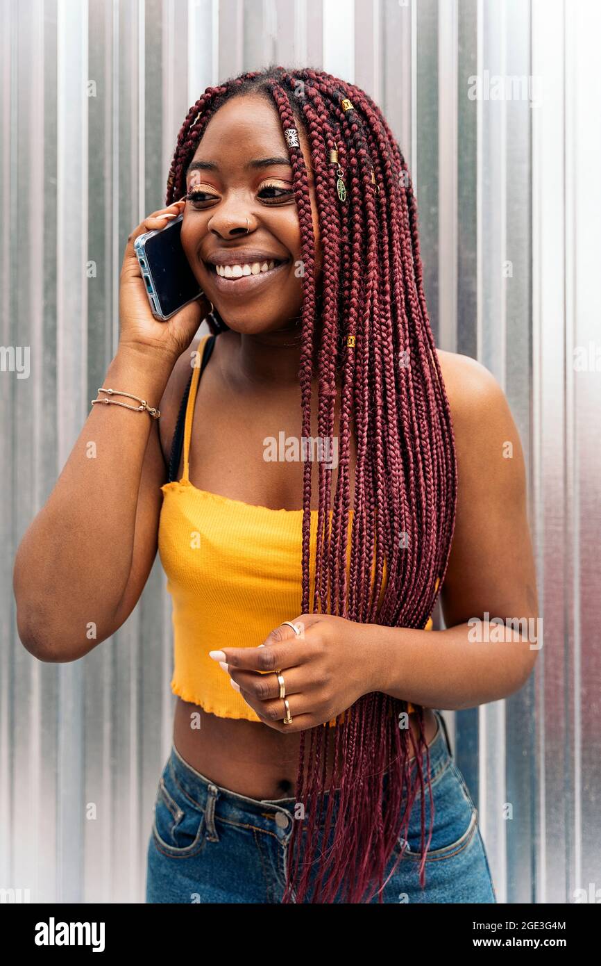 Fröhliche junge afrikanische Frau mit Zöpfen, die auf der Straße stehen und am Telefon sprechen. Stockfoto