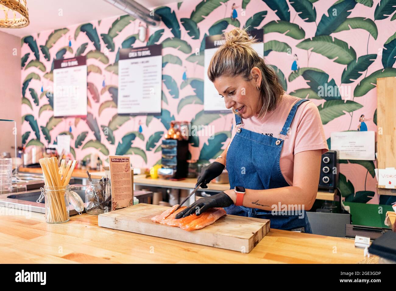Eine Restaurantarbeiterin trägt eine Schürze und Latexhandschuhe und schneidet Lachs für eine Poke-Schüssel. Stockfoto