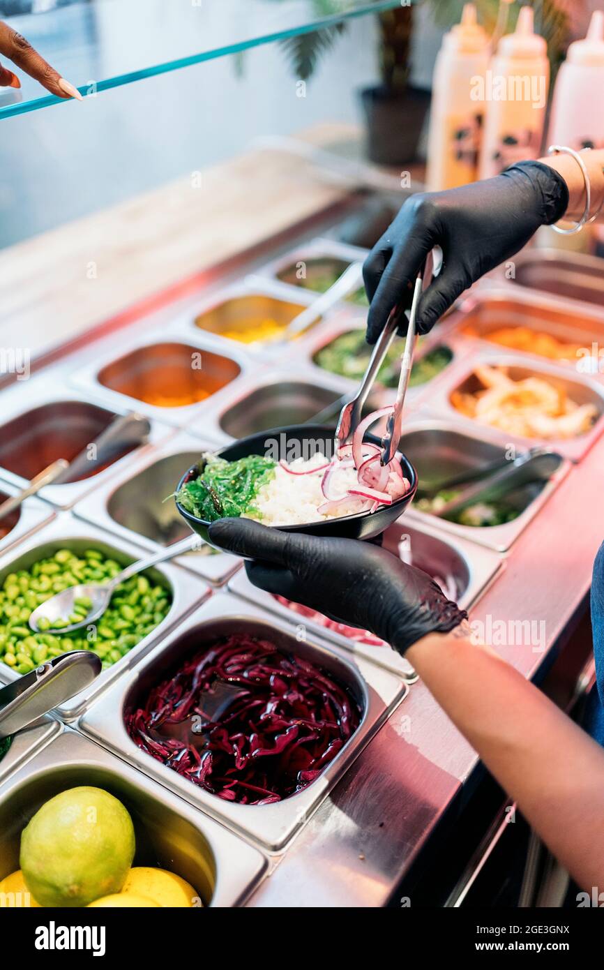 Nicht anerkannter Restaurantmitarbeiter mit Schutzhandschuhen, der Zutaten in eine Schüssel mit Reis und Gemüse steckt. Stockfoto