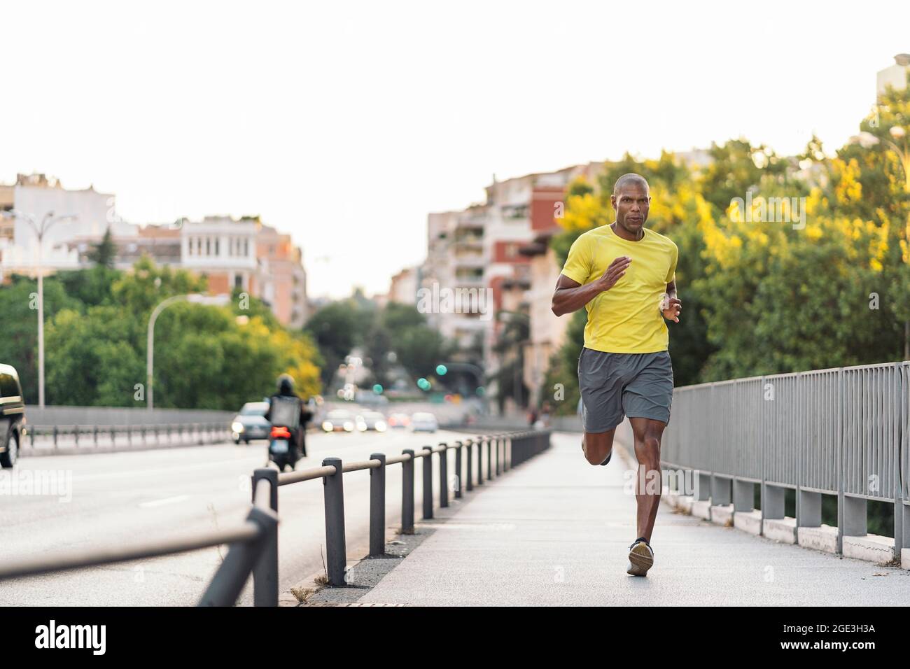 Vorderansicht eines afroamerikanischen Sportlers mit gelbem T-Shirt und Shorts beim Joggen auf dem Bürgersteig in der Stadt im Freien. Stockfoto