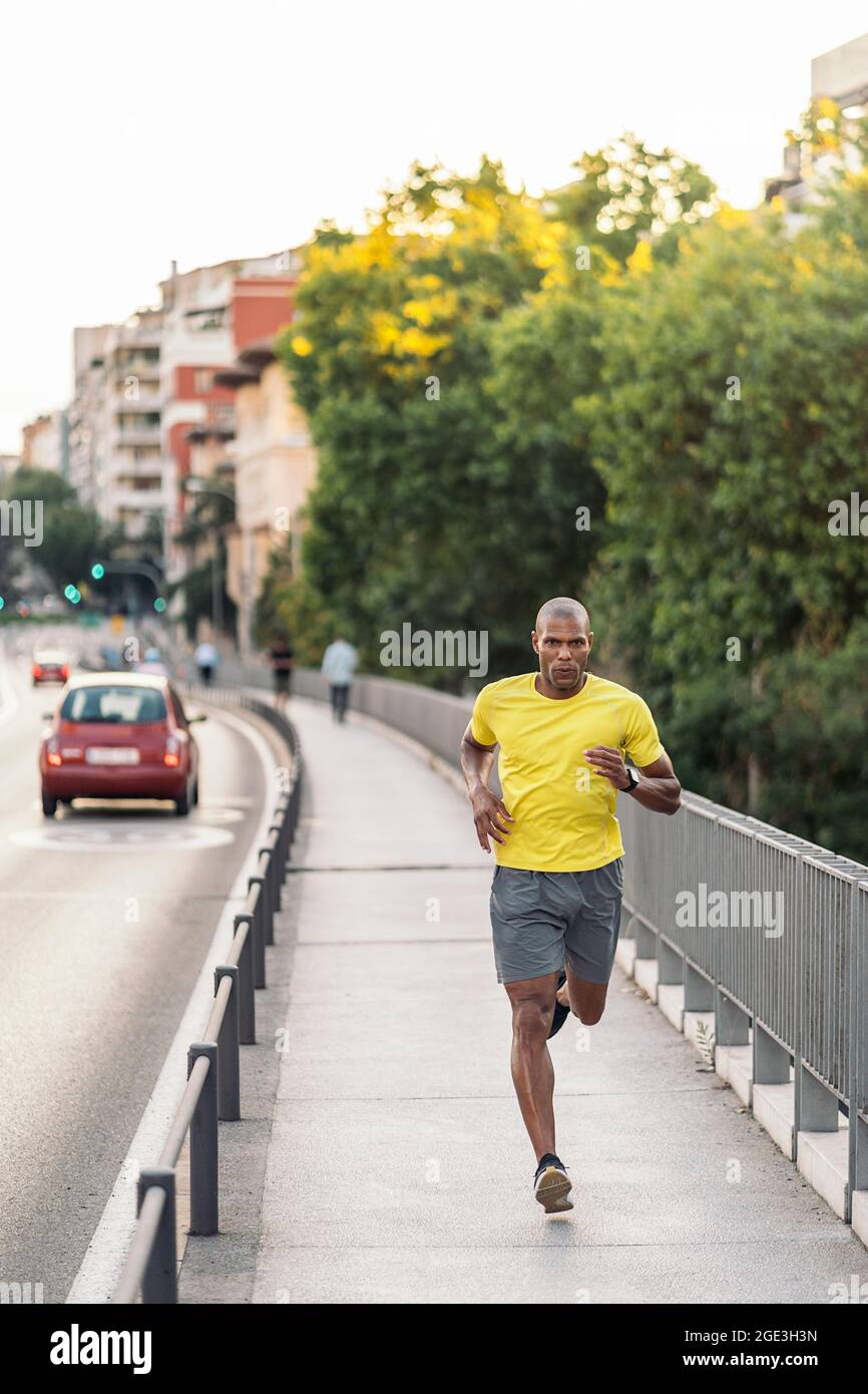 Vorderansicht eines afroamerikanischen Sportlers mit gelbem T-Shirt und Shorts beim Joggen auf dem Bürgersteig in der Stadt im Freien. Stockfoto