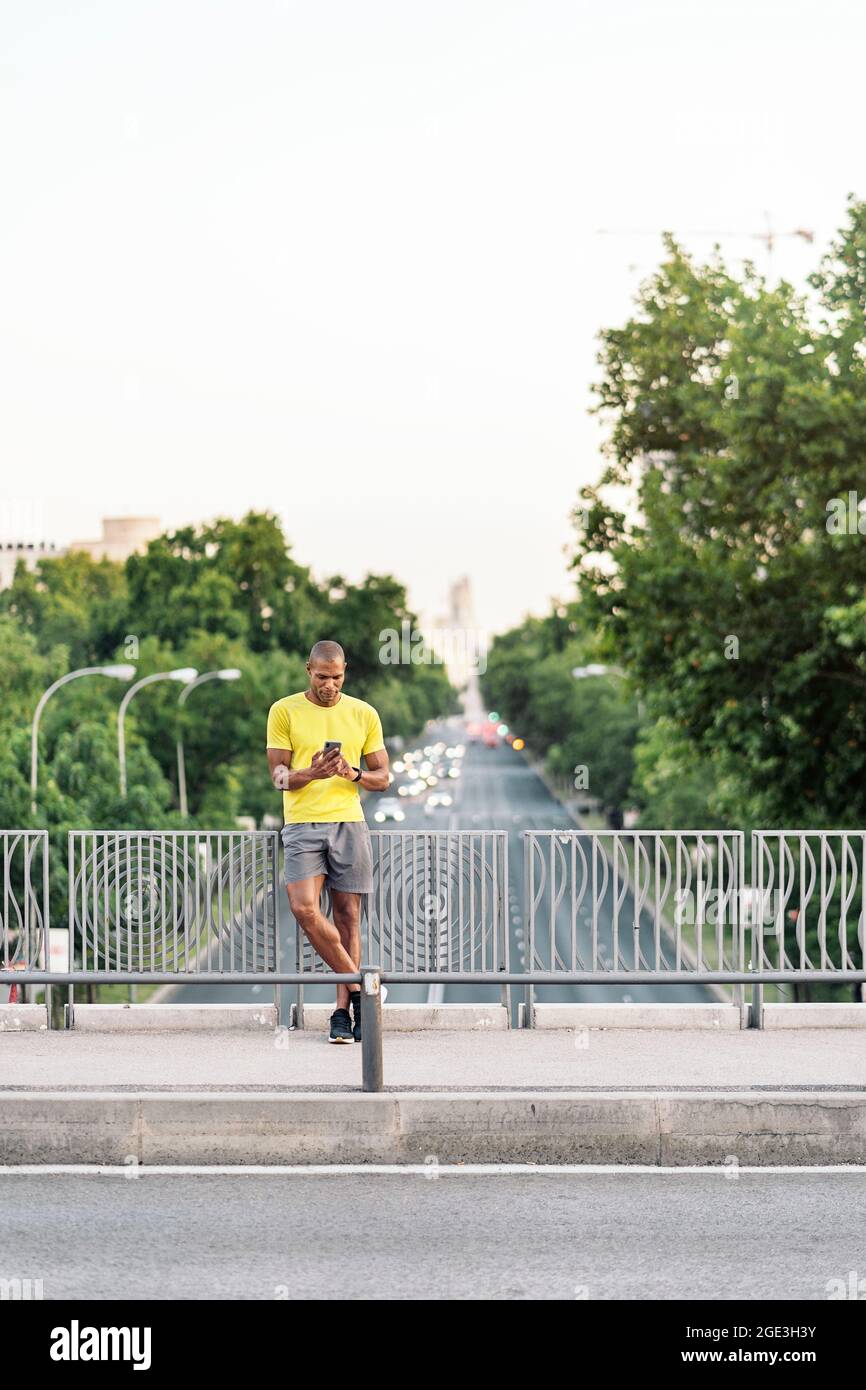 Vorderansicht eines afroamerikanischen Sportlers mit gelbem T-Shirt und Shorts beim Joggen auf dem Bürgersteig mit dem Telefon in einer Fußgängerbrücke im Freien. Stockfoto