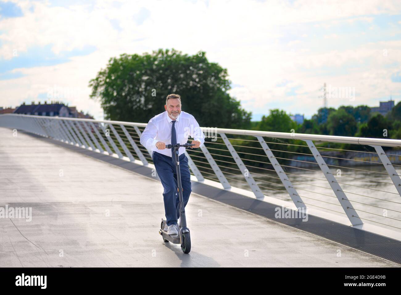 Ein lustiger Geschäftsmann genießt seine Fahrt mit einem Elektroroller, als er bei Sonnenuntergang eine lange Brücke überquert und dabei den Fußgängerweg in einem städtischen Nahverkehr benutzt Stockfoto