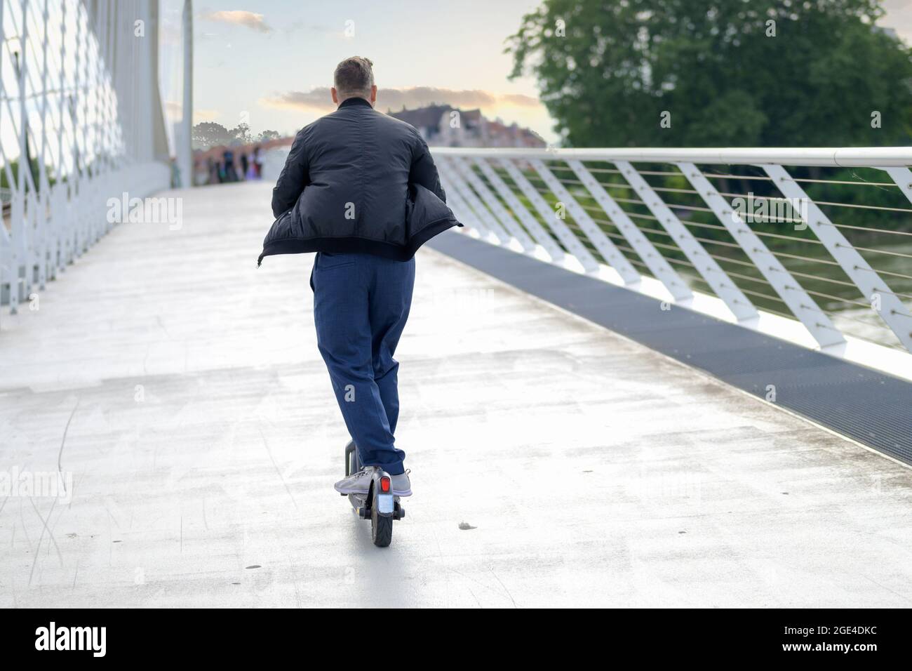 Rückansicht eines Mannes, der bei Sonnenuntergang auf einer Brücke auf einen Elektroroller reitet, auf weit entfernte Menschen, die in einem städtischen Transportkonzept den Weg überqueren Stockfoto