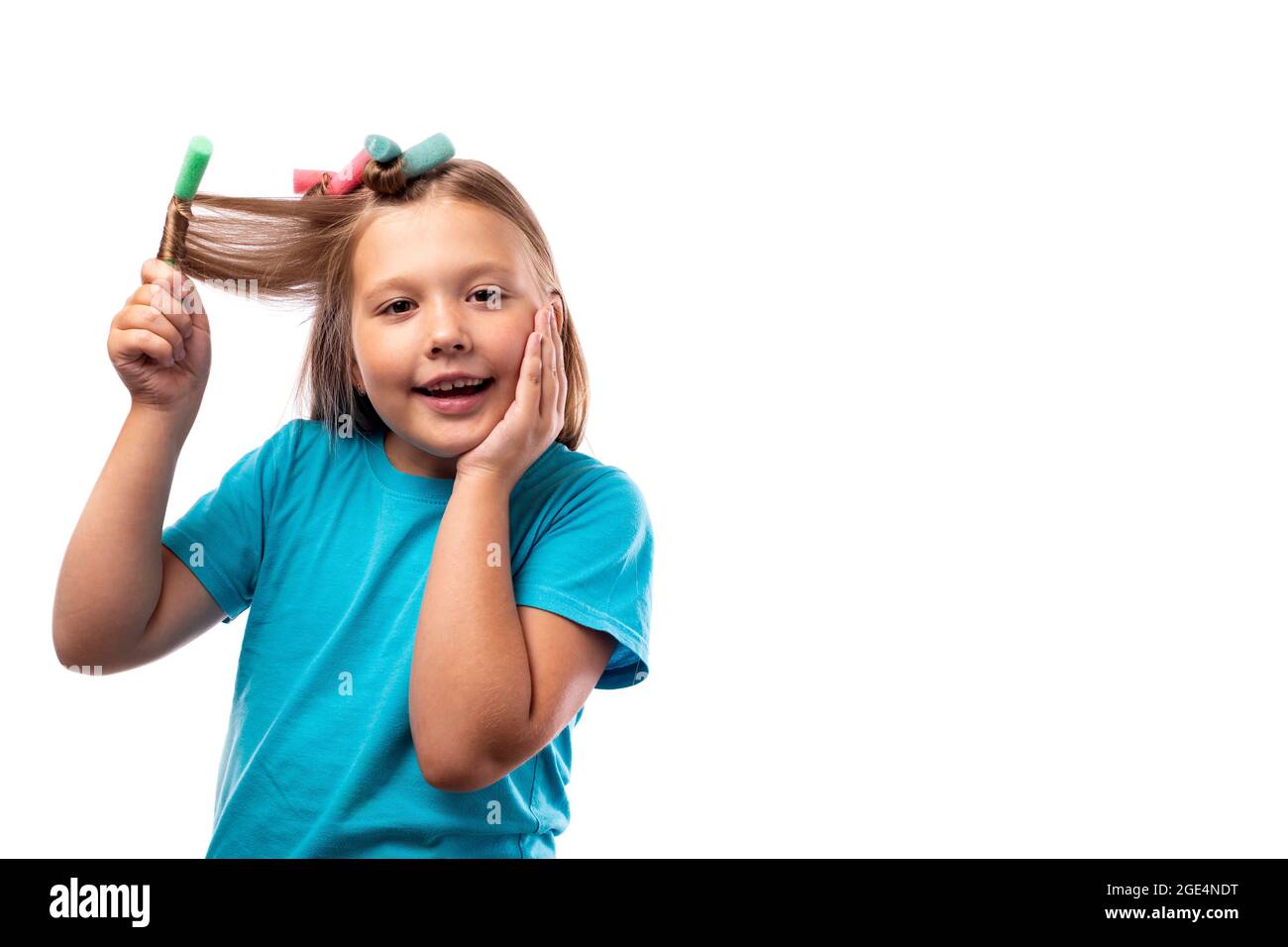 Ein glückliches kaukasisches kleines Mädchen in einem blauen T-Shirt wickelt ihr langes blondes Haar in Lockenwickler auf einem hellen Hintergrund. Lockern Sie Ihr Haar mit mehrfarbigen Lockenwicklern. Stockfoto