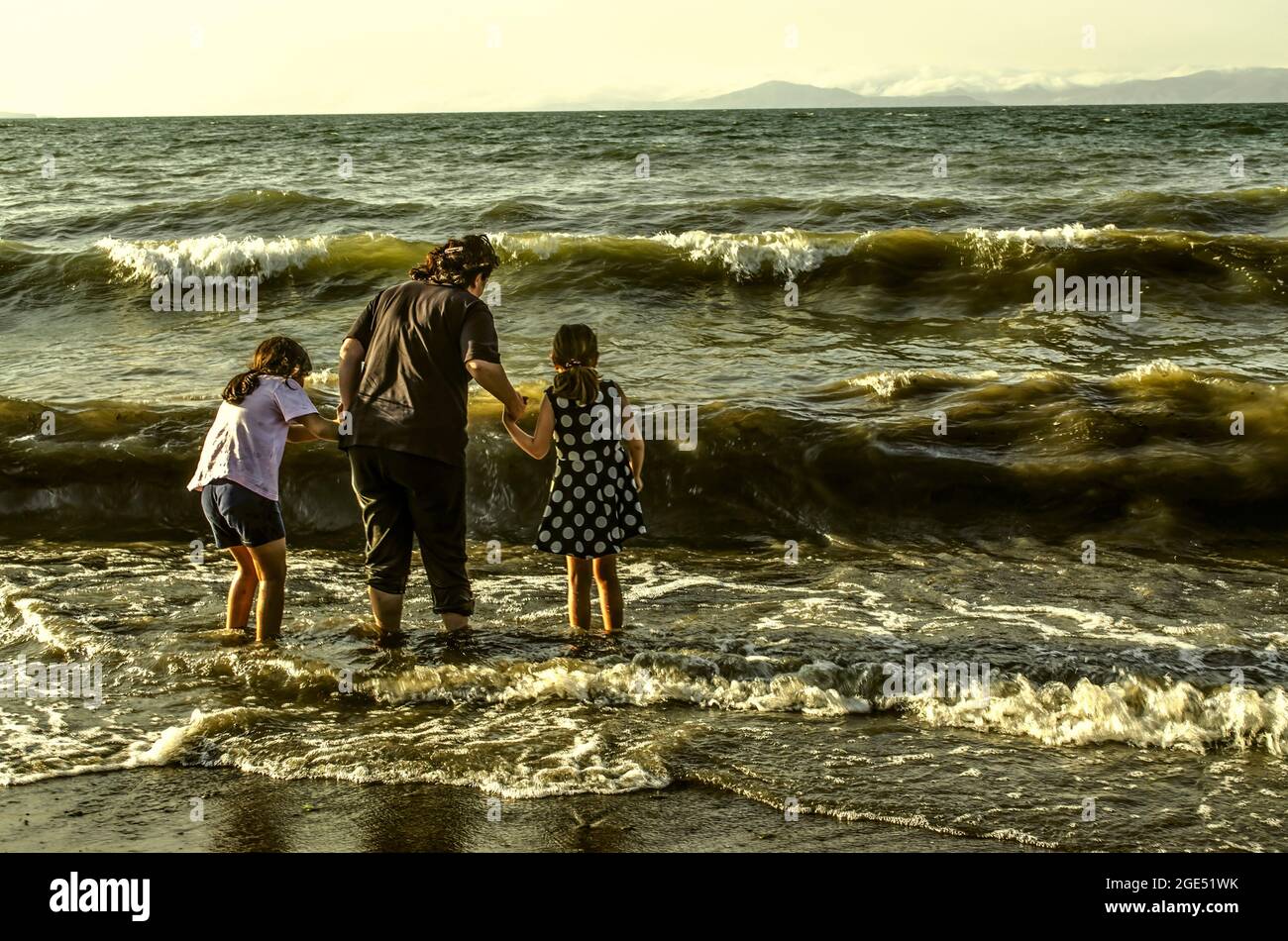 Sevan-See, Armenien, 24. Juni 2021: Eine junge Frau hält zwei kleine Mädchen an den Händen und steht knietief im stürmischen, schaumigen Wasser, beleuchtet von der r Stockfoto