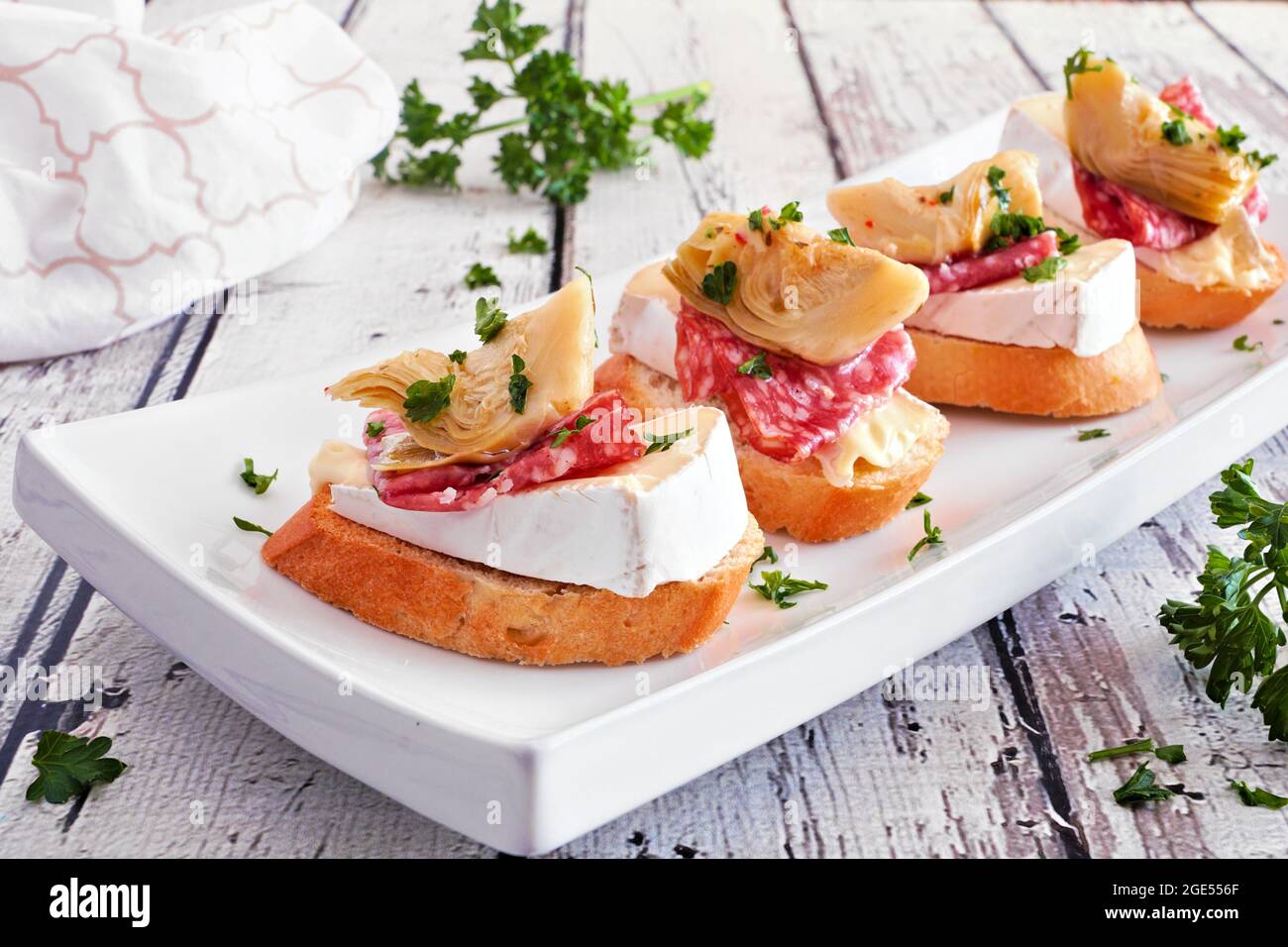 Crostini-Vorspeisen mit Brie-Käse, Salami und Artischocken. Nahaufnahme auf einem Servierteller vor einem weißen Holzhintergrund. Party-Food-Konzept. Stockfoto