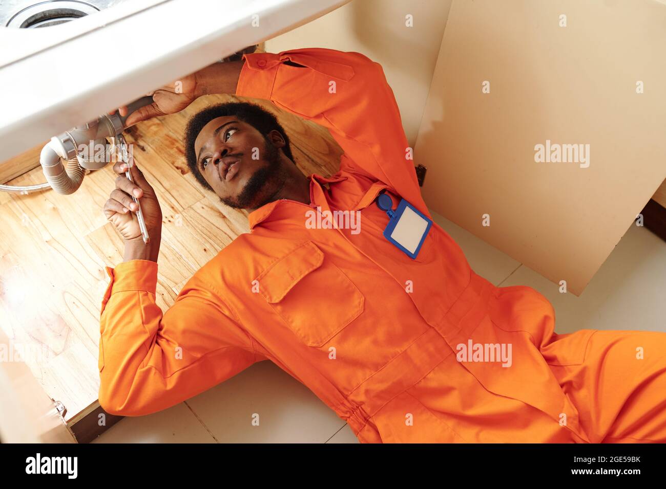 Junger Klempner in orangefarbener Uniform, der unter dem Waschbecken liegt und undichte Rohre fixiert Stockfoto