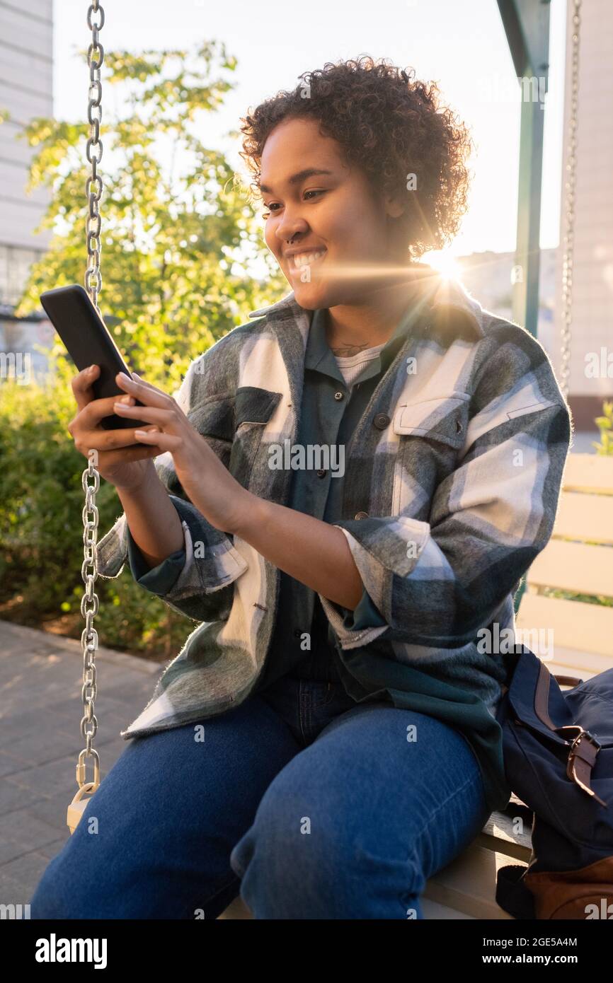 Glückliche afrikanische weibliche Teenager mit Smartphone auf Schaukel in der städtischen Umgebung sitzen Stockfoto