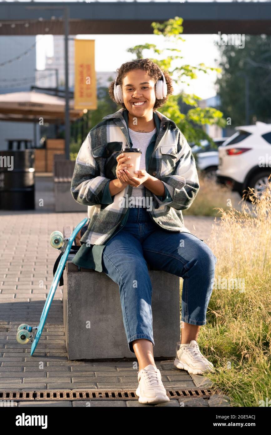 Fröhliche Teenager-Skateboarderin mit Kopfhörern, die im Park einen Drink zu sich nehmen Stockfoto