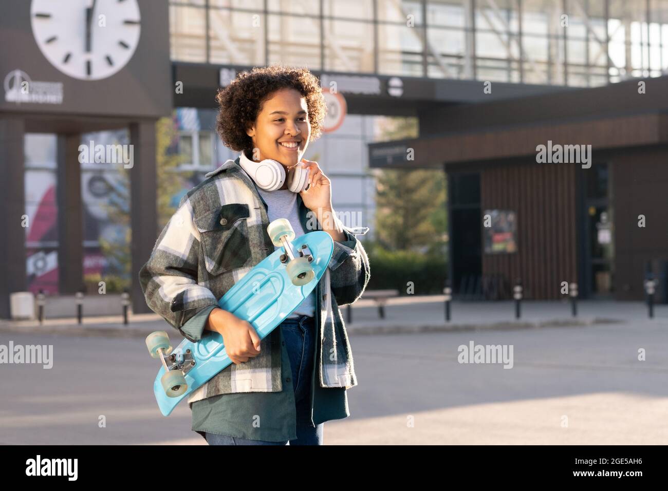 Lächelnde Teenager mit blauem Skateboard, während sie gegen moderne Architektur steht Stockfoto