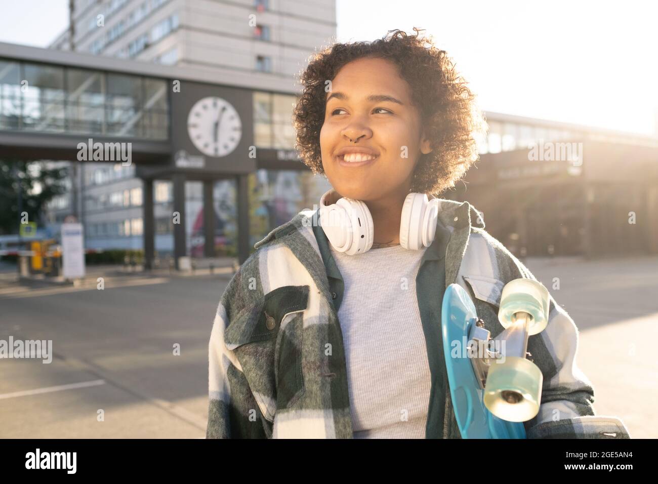 Netter, fröhlicher Teenager mit Skateboard und Kopfhörern, die im urbanen Umfeld stehen Stockfoto