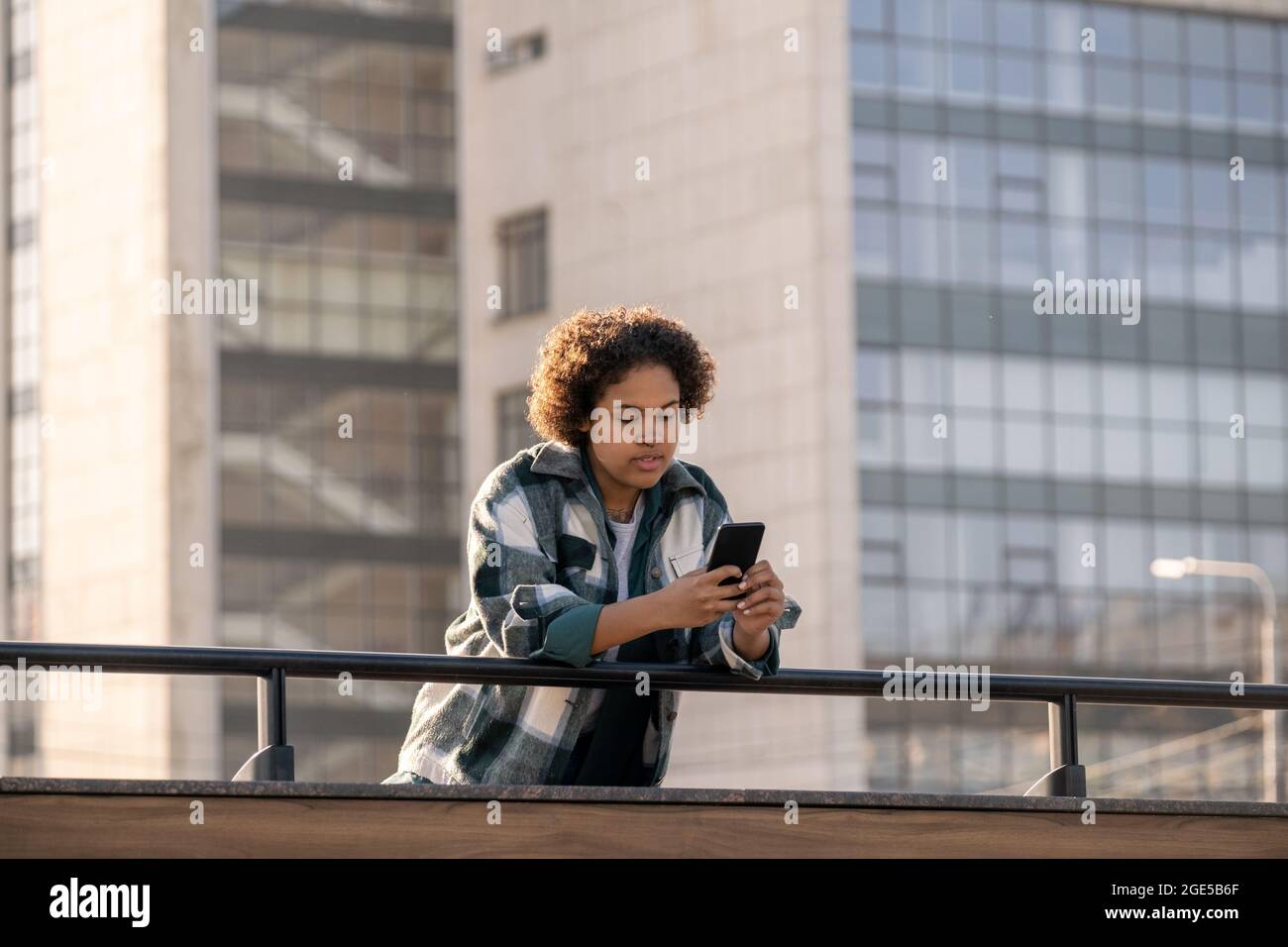 Weibliche Teenager scrollt im Smartphone, während sie sich gegen moderne Architektur über Geländer beugt Stockfoto