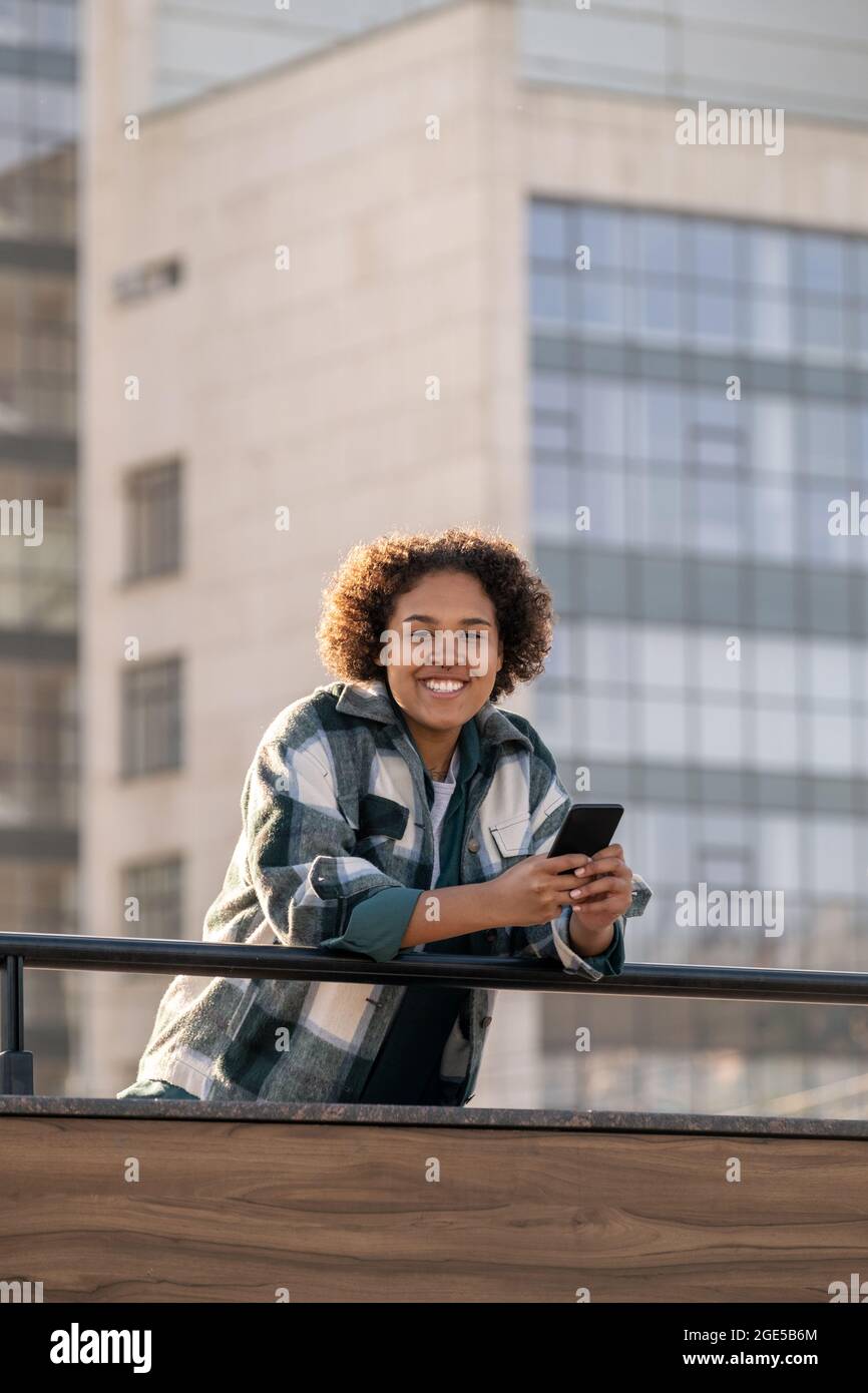 Ein hübscher Teenager, der Sie mit toothy Lächeln ansieht, während er im Smartphone scrollt Stockfoto