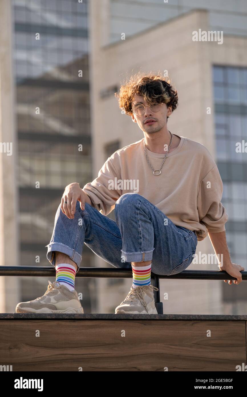 Moderner Teenager-Typ in Jeans und Sweatshirt, der auf Geländern an der Außenfassade des Gebäudes sitzt Stockfoto