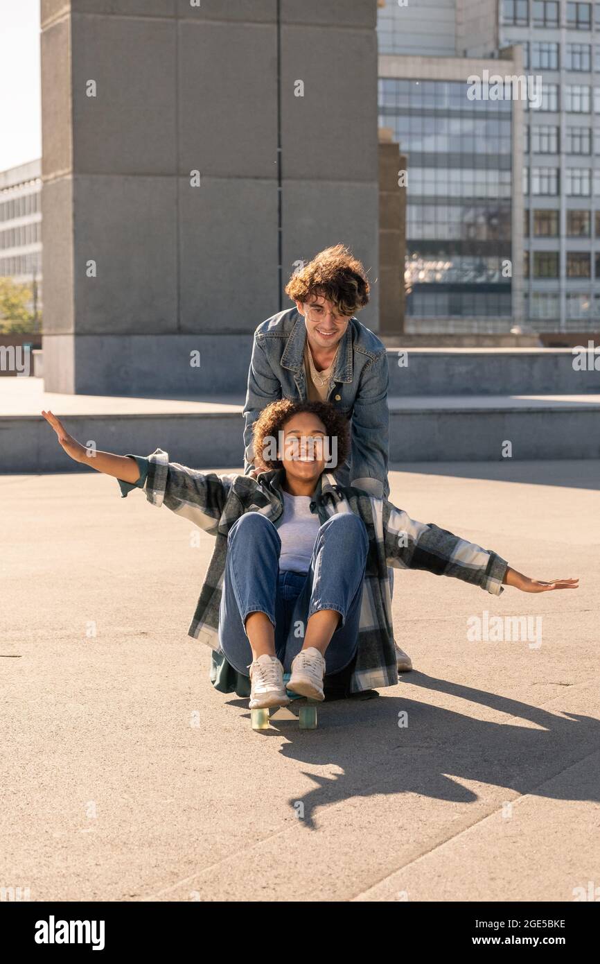 Fröhlicher Teenager, der seine afrikanische Freundin auf Skateboard schiebt, während beide Spaß haben Stockfoto
