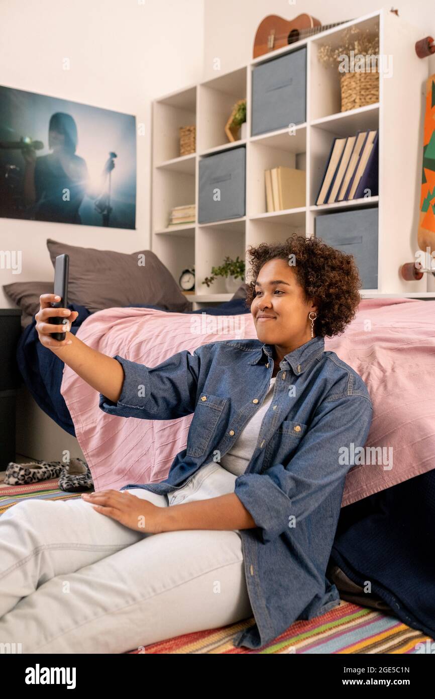 Glücklicher afrikanischer Teenager mit Smartphone, das im Video-Chat kommuniziert, während er am Bett auf dem Boden sitzt Stockfoto