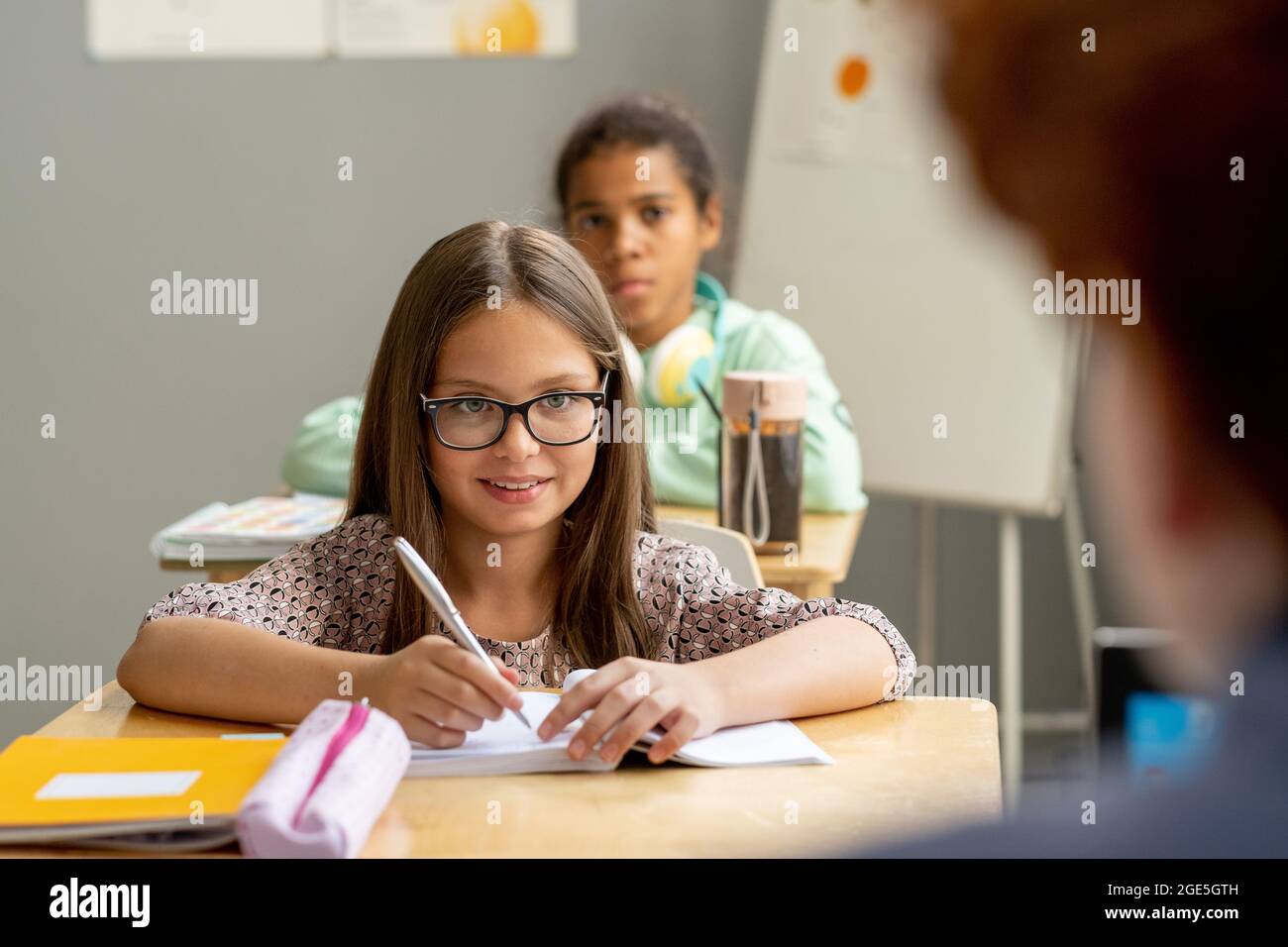 Glückliche Schülerin in Brillen Blick auf Lehrer, während Notizen in Copybook in der Lektion Stockfoto